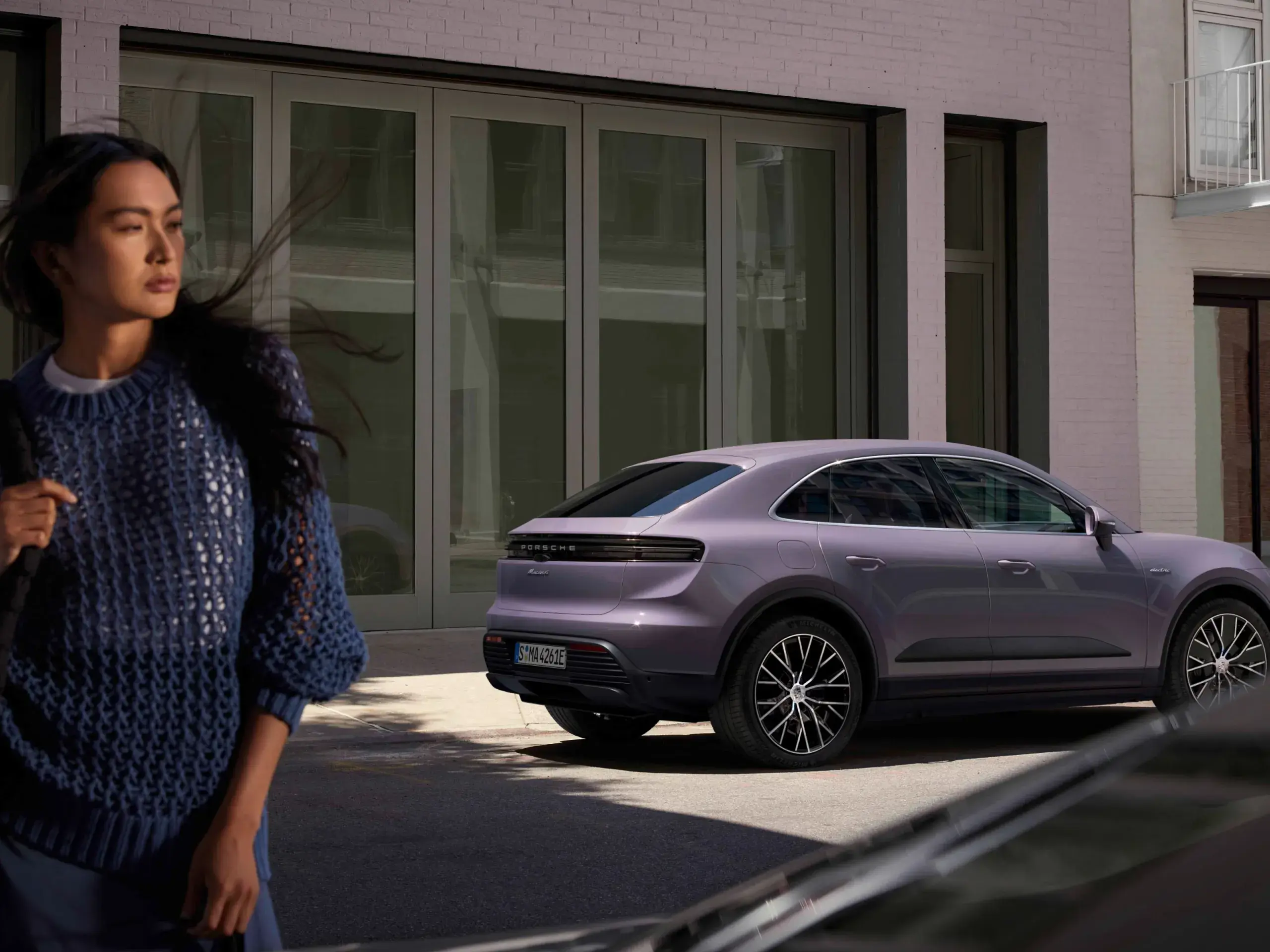 En kvinde går forbi en parkeret lilla Porsche Macan foran en moderne bygning i byen.