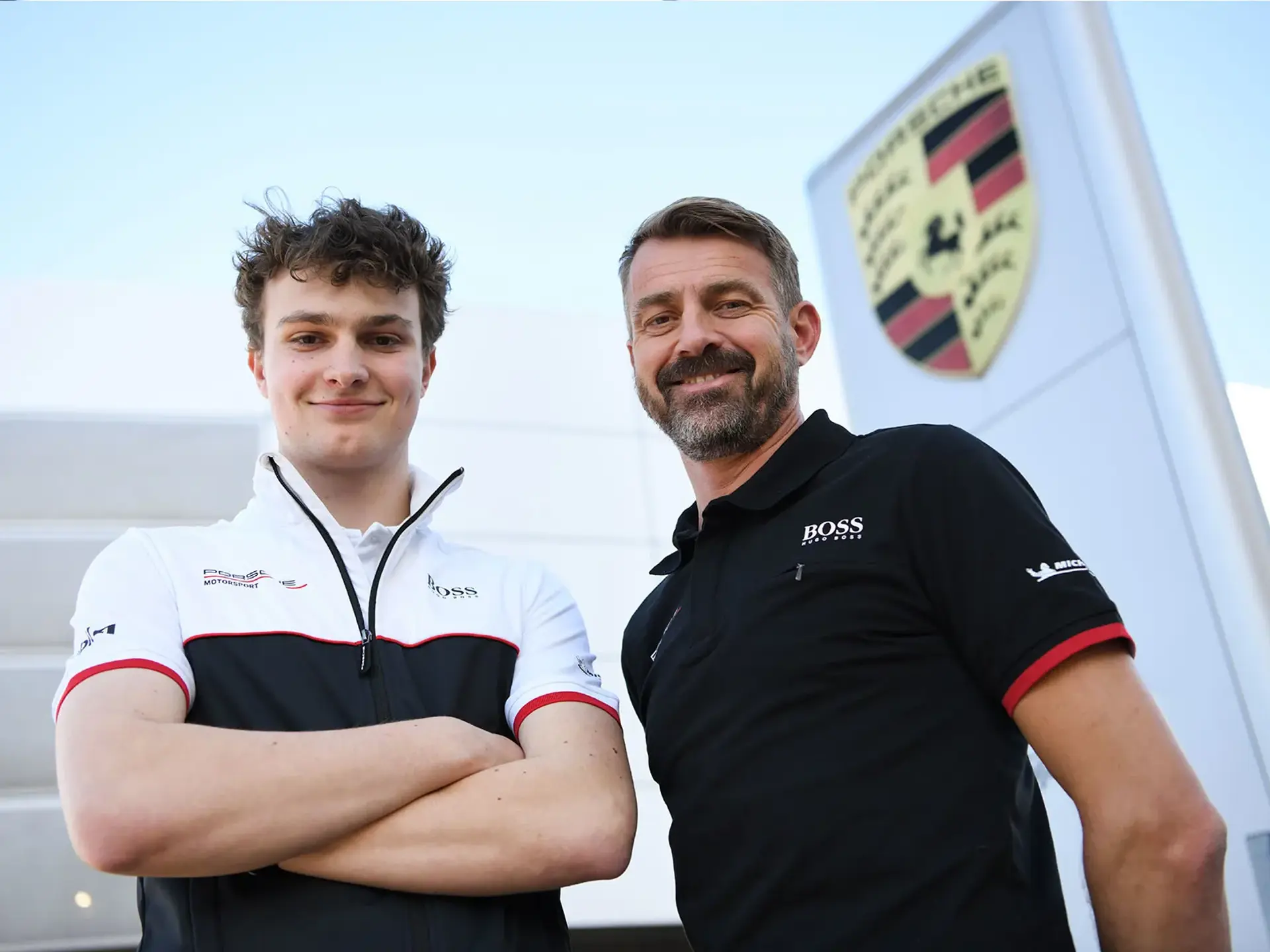 To Porsche Denmark-racerkørere står foran et stort Porsche-logo ved et moderne anlæg.