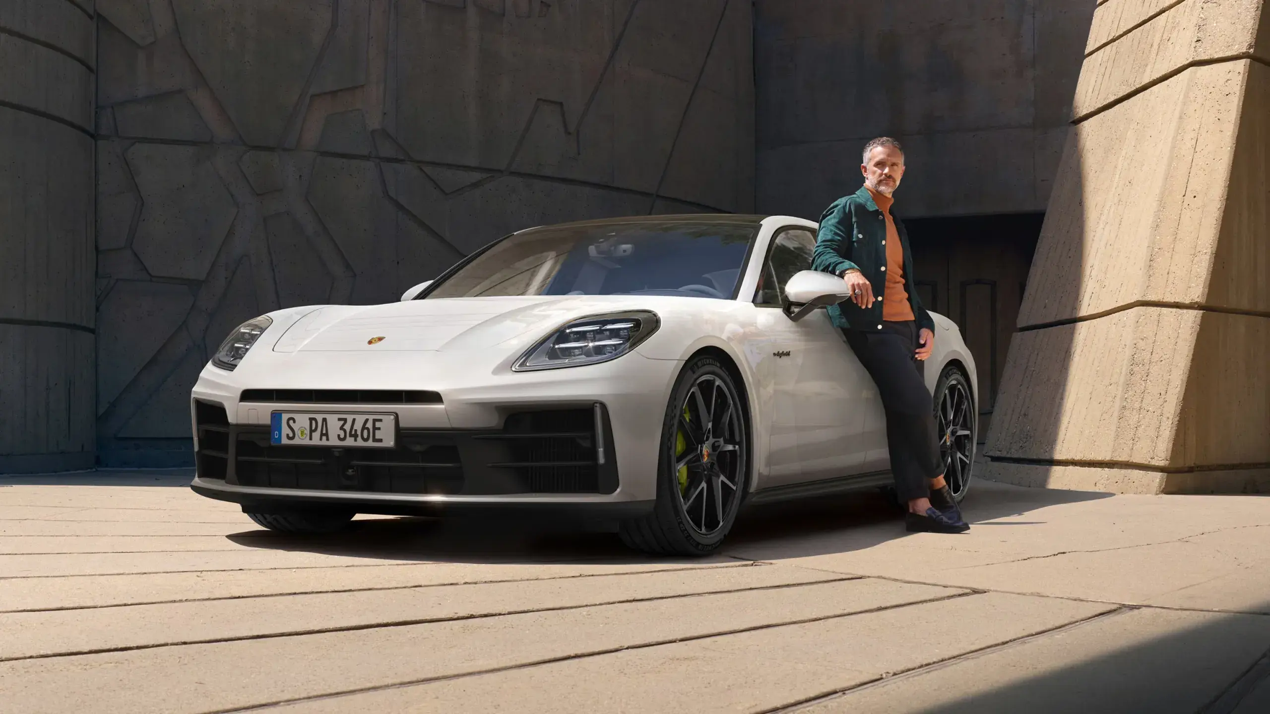 En mand læner sig op ad en hvid Porsche Panamera 4 E-Hybrid parkeret foran en moderne betonbygning.