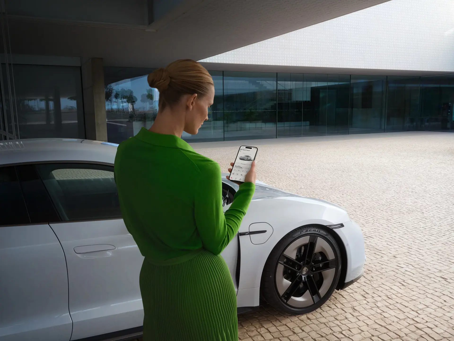 Kvinde i grønt står ved en hvid Porsche Taycan og styrer bilen via en app på sin smartphone.