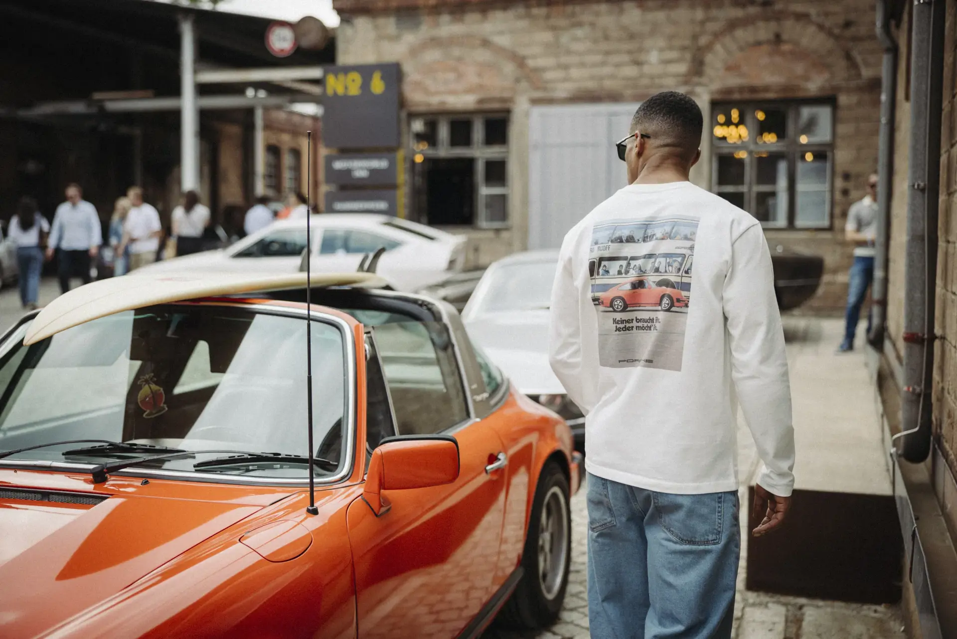 Mand i casual tøj går forbi en orange klassisk Porsche parkeret i et urbant miljø med mennesker i baggrunden.