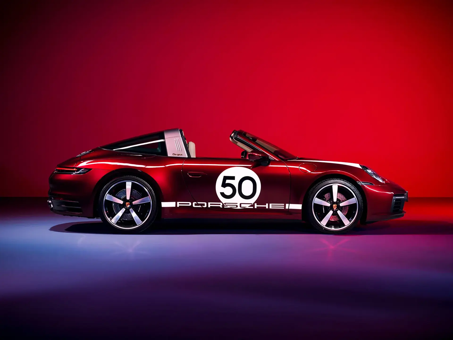 Rød Porsche 911 cabriolet i heritage-design med startnummer 50 set fra siden mod en rød baggrund.