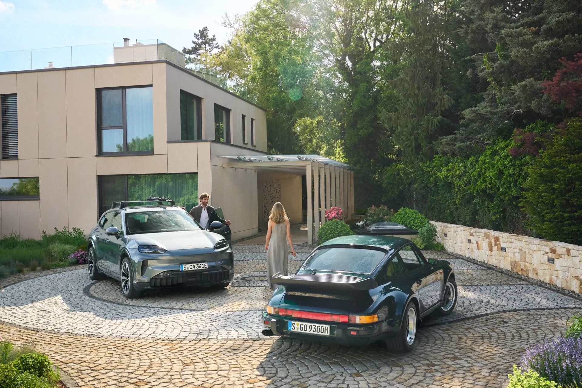En mand og en kvinde står i indkørslen foran en moderne villa mellem en elektrisk Porsche Cayenne og en klassisk Porsche sportsvogn.