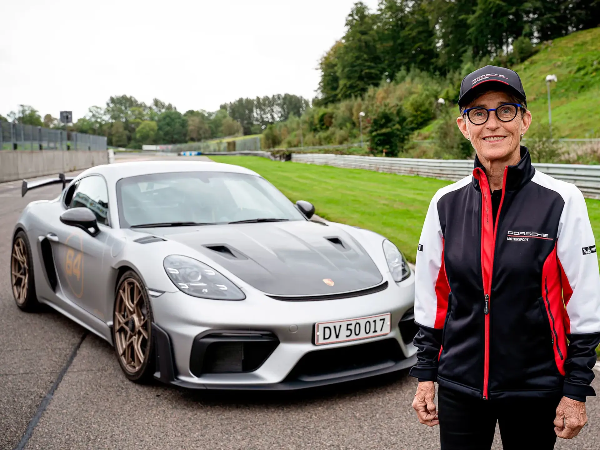 Person i Porsche Motorsport-tøj står foran en sølvgrå Porsche sportsvogn på en racerbane.