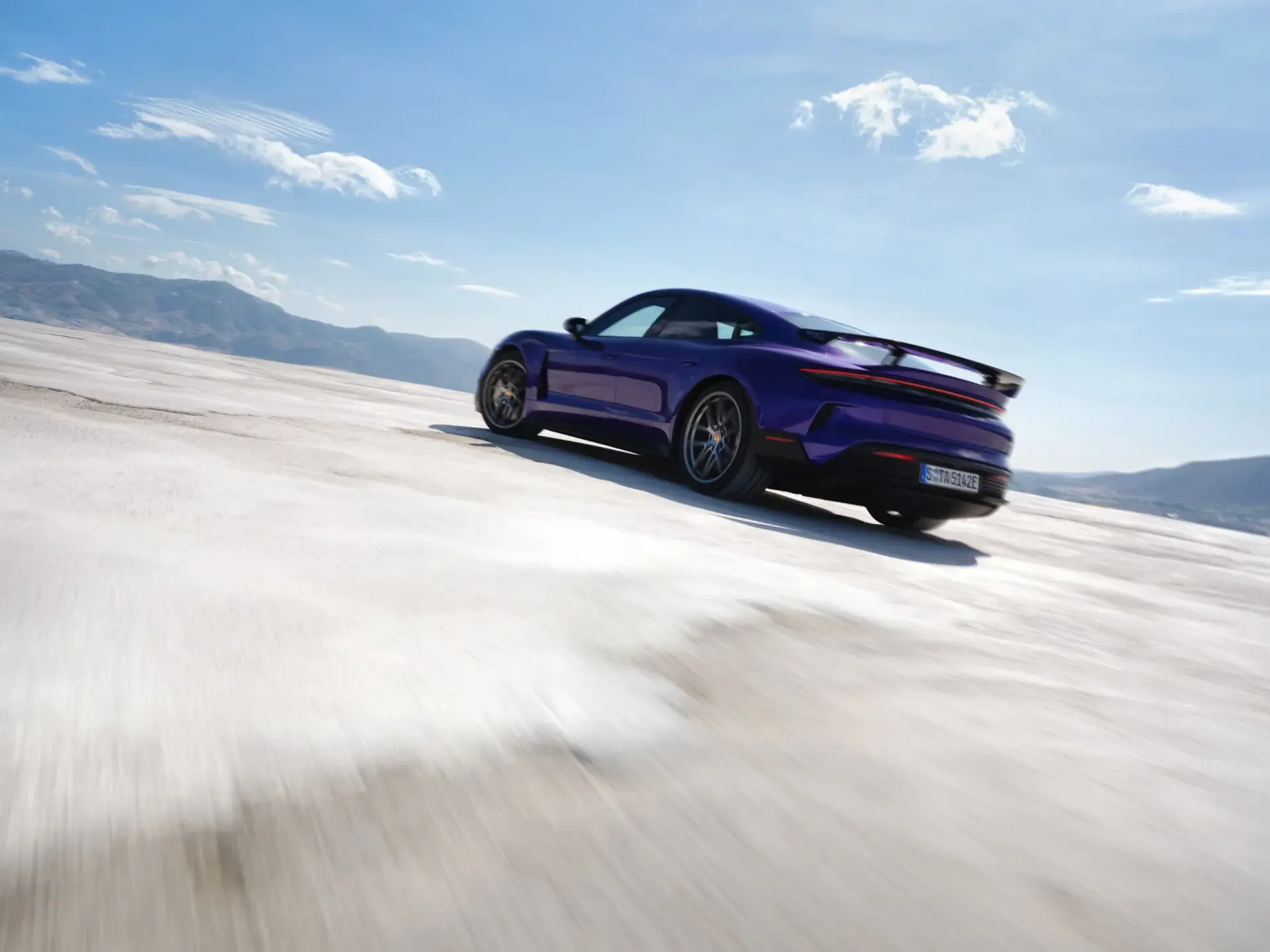 En blå Porsche Taycan Turbo GT med Weissach-pakke kører hurtigt hen over et åbent, lyst landskab med bjerge i baggrunden.