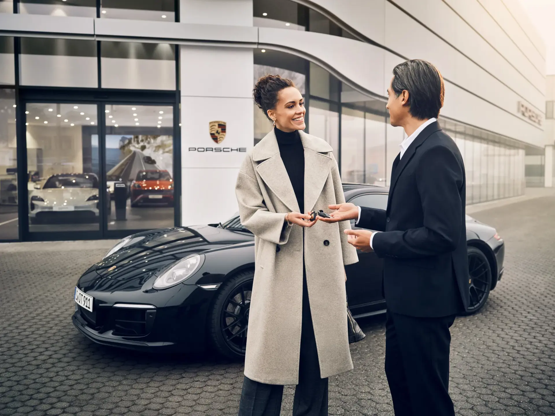 En smilende kunde modtager nøglerne til en sort Porsche foran et Porsche-center.