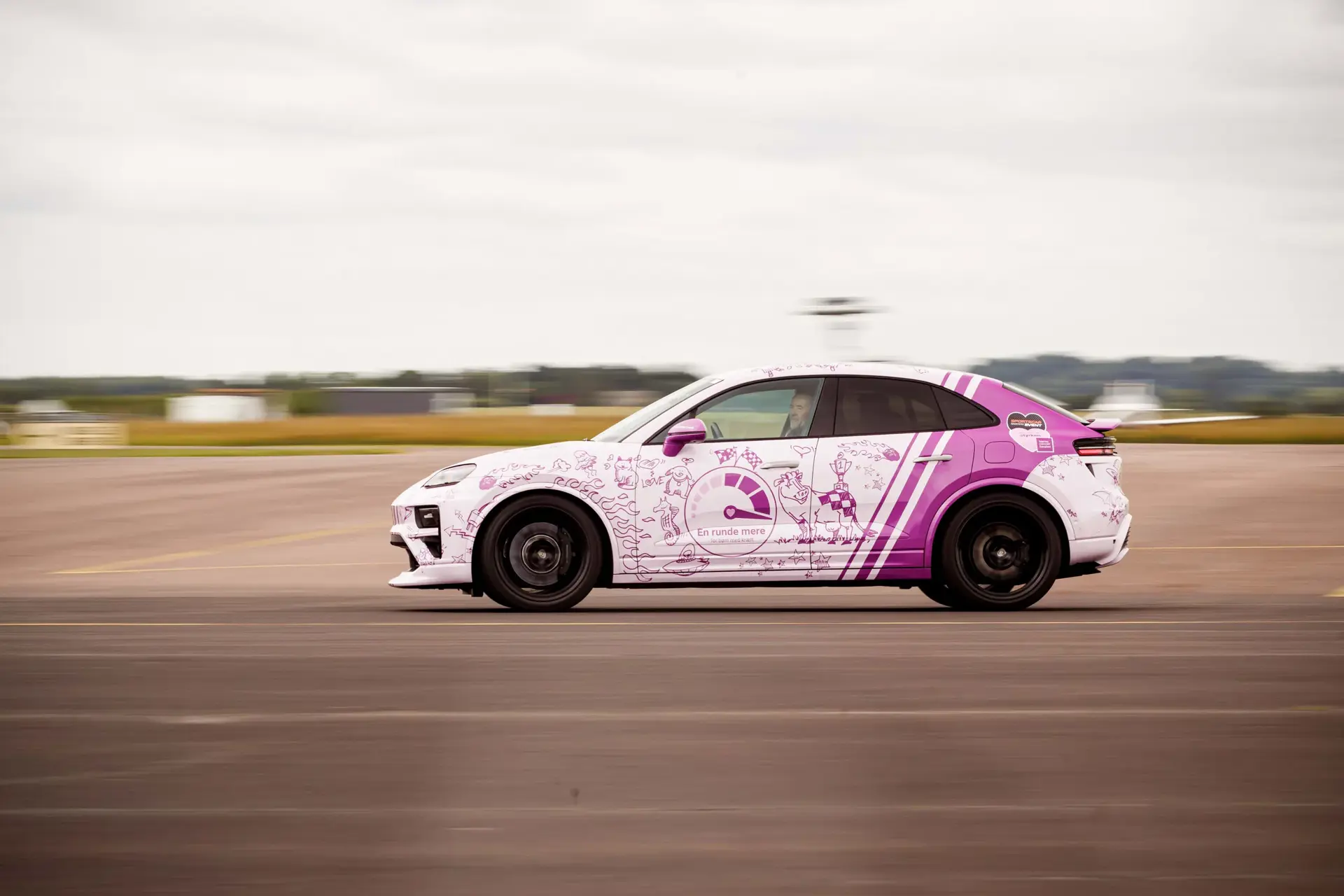 Pink og hvid sportsbil med børnetegninger og teksten "En runde mere" kører hurtigt på en racerbane.