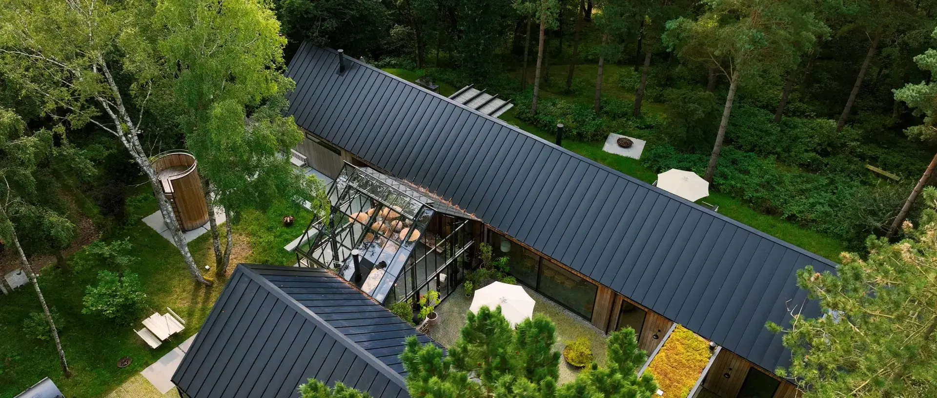 Luftfoto af The Barnhouse Project, et moderne træhus med glasudestue omgivet af tæt grøn skov og havearealer.