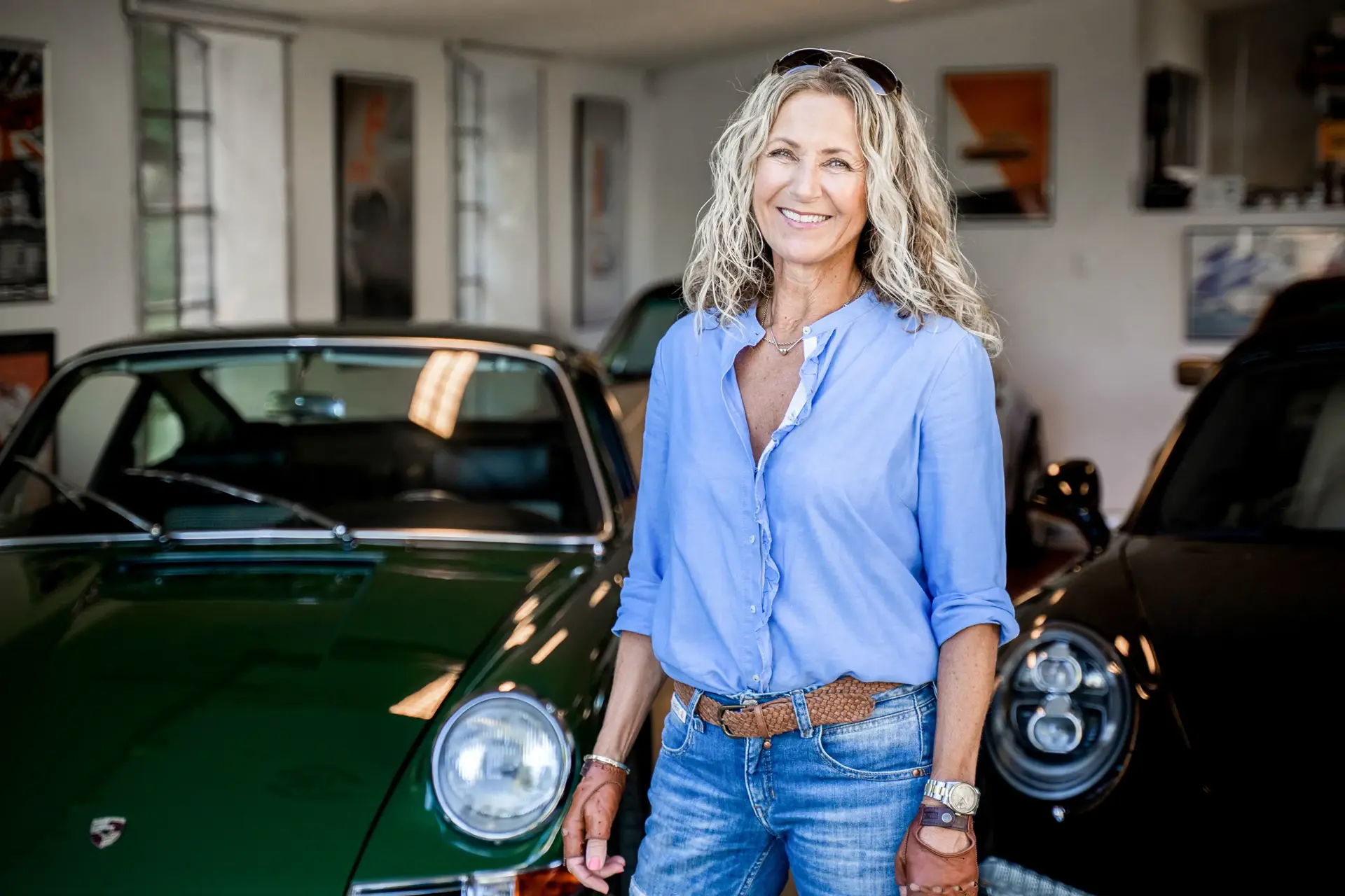 Smilende kvinde i lyseblå skjorte står i en garage mellem to klassiske Porsche-biler.