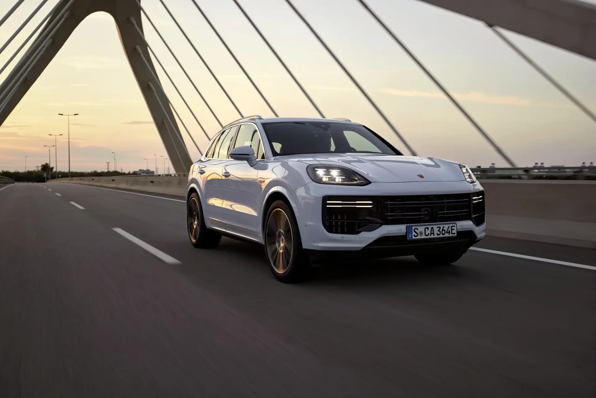 Hvid Porsche SUV kører over en moderne bro ved solnedgang.