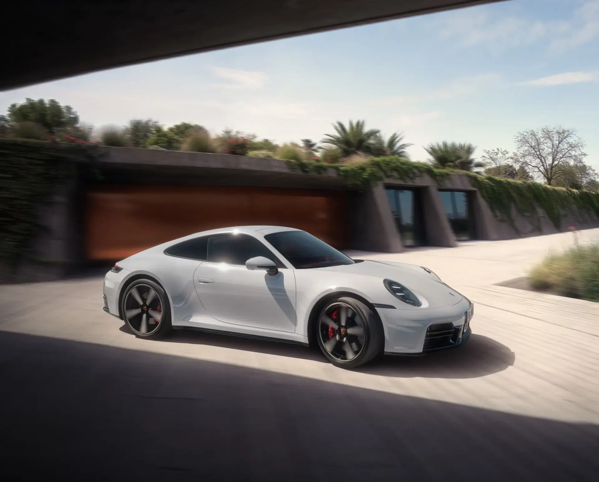 En hvid Porsche 911 Carrera S kører i høj fart forbi en moderne bygning omgivet af grøn beplantning.