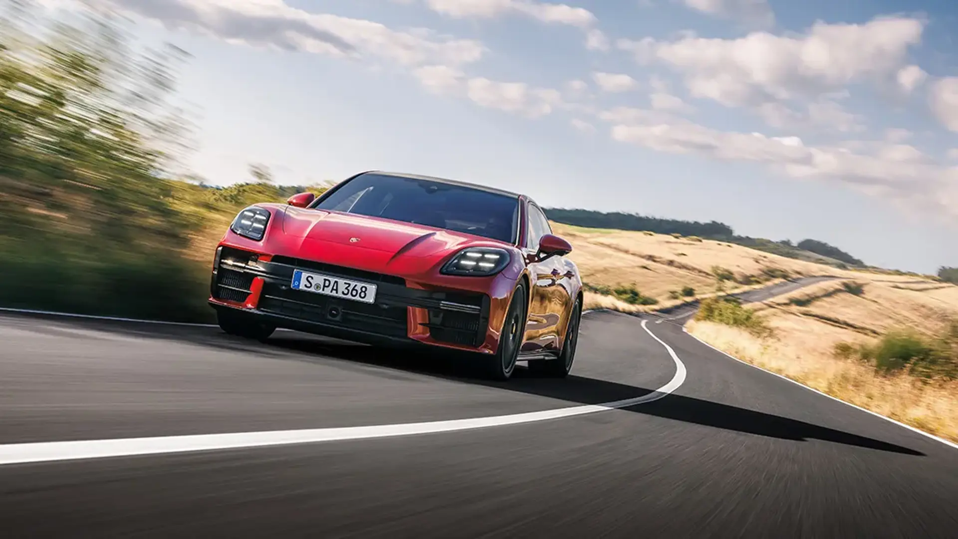 Rød Porsche Panamera ses forfra, mens den kører hurtigt ad en snoet landevej gennem et åbent, solrigt landskab.