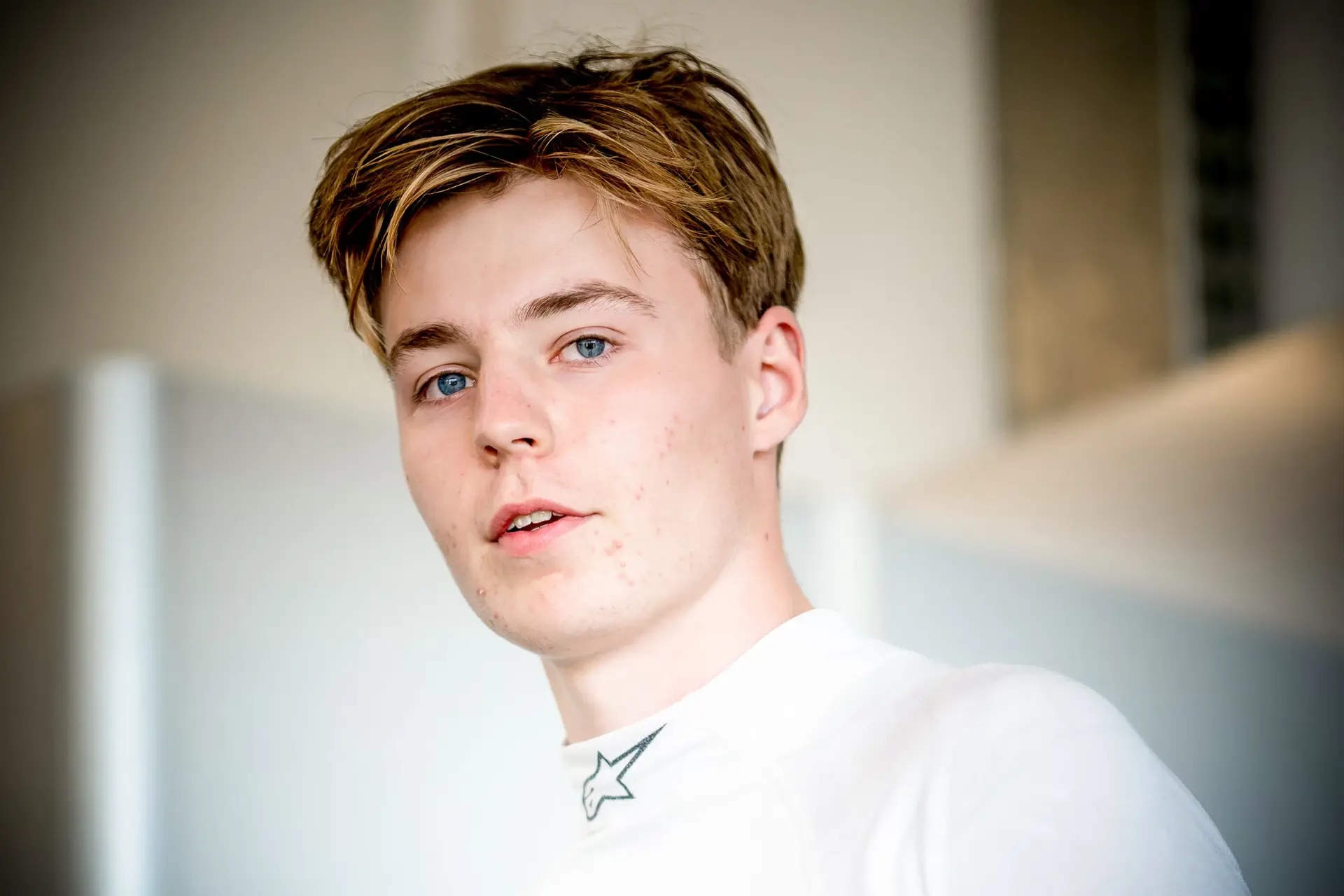 Ung mand i hvid køredragt med Alpinestars-logo står indendørs i blødt lys.