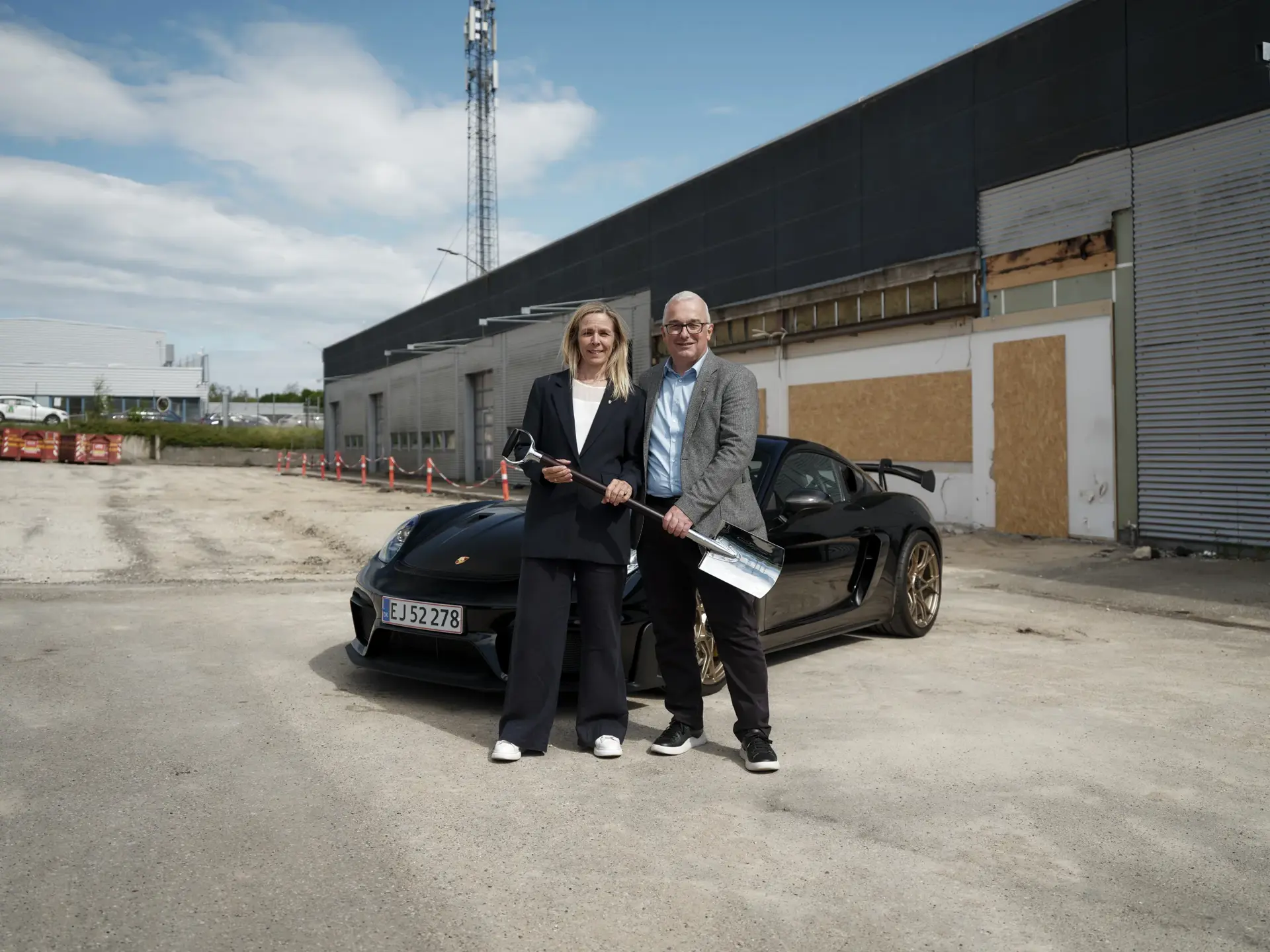 To personer med ceremonielle spader står foran en sort sportsvogn på en byggeplads ved en industribygning.