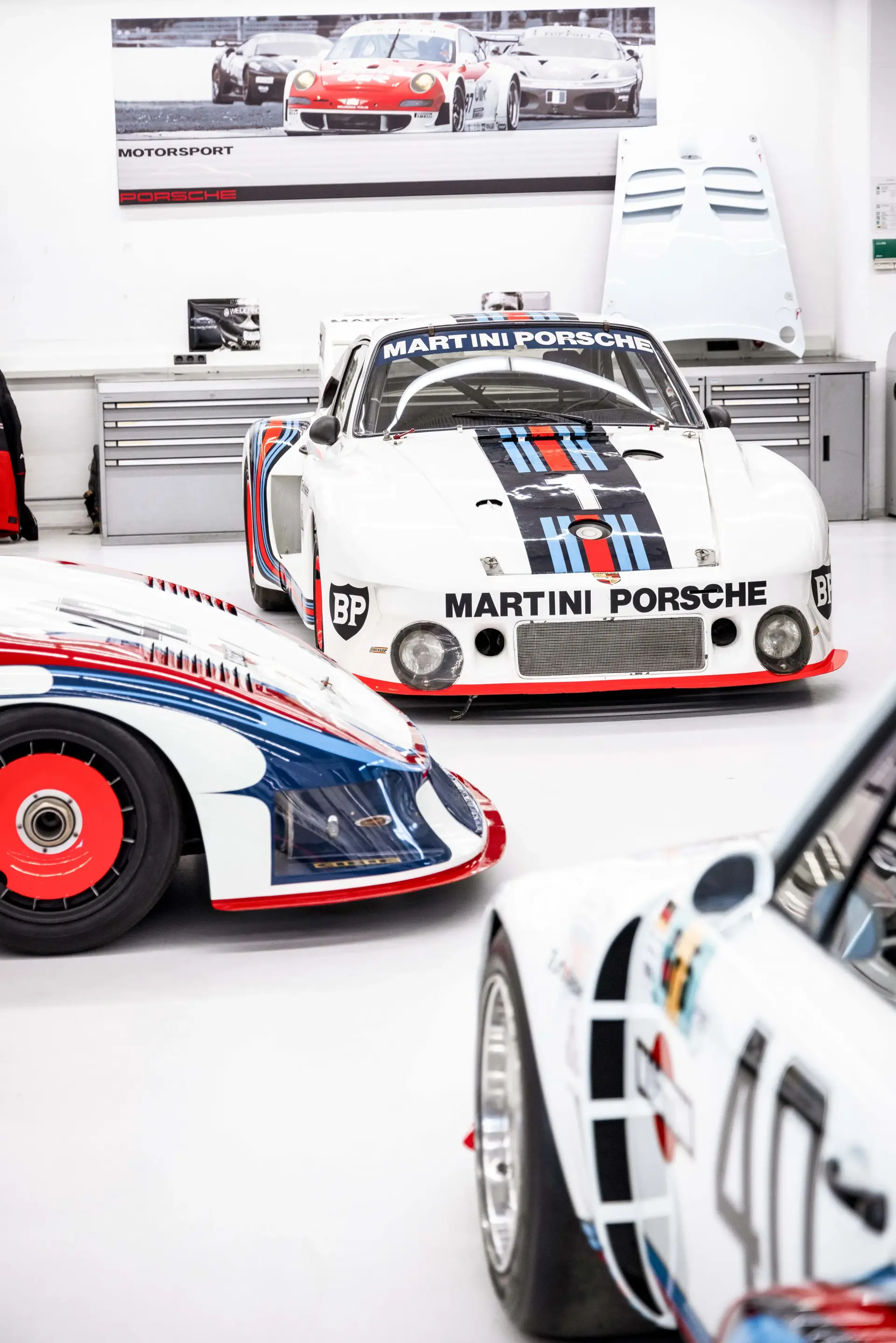 Flere klassiske Porsche-racere i Martini-bemaling står udstillet i et lyst værksted.