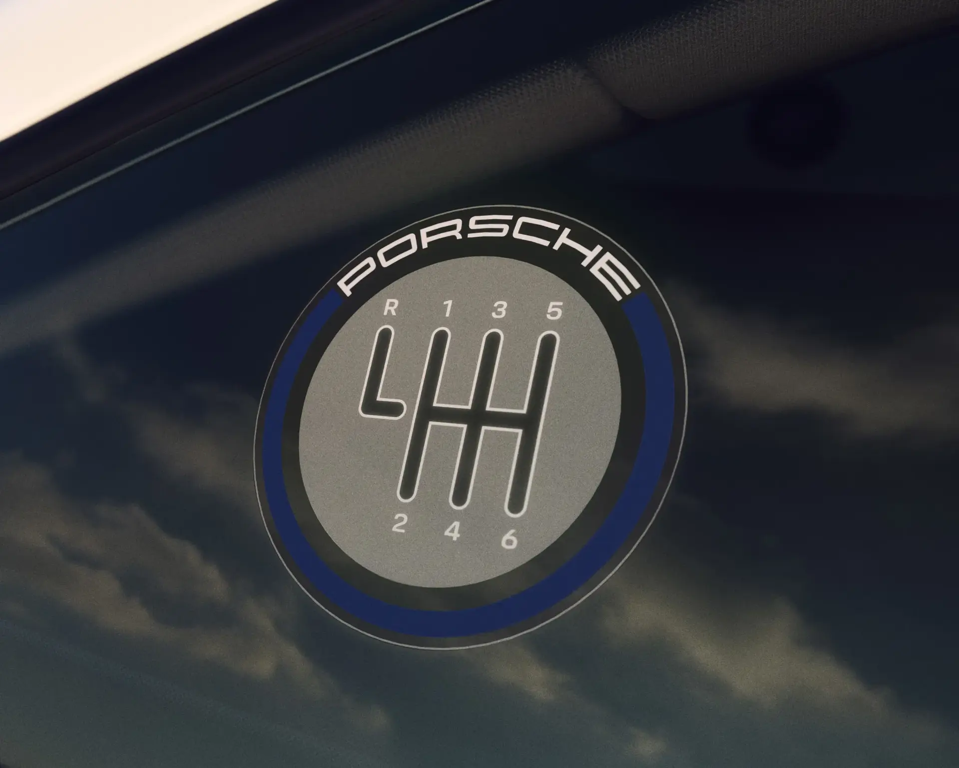 Rundt Porsche-emblem på en bilrude med illustration af et manuelt gearskema.