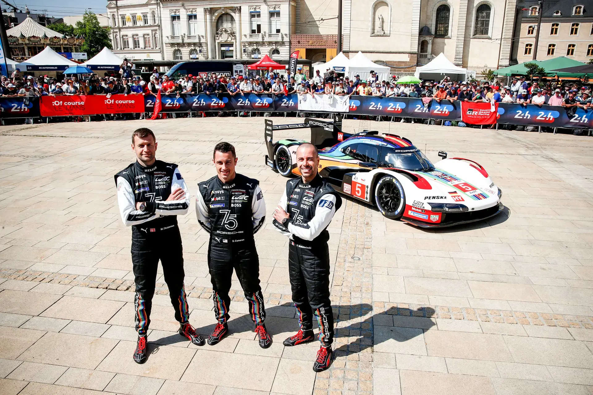 Tre racerkørere i Porsche-kørerdragter poserer foran en Porsche Le Mans-racerbil på en plads med publikum i baggrunden.