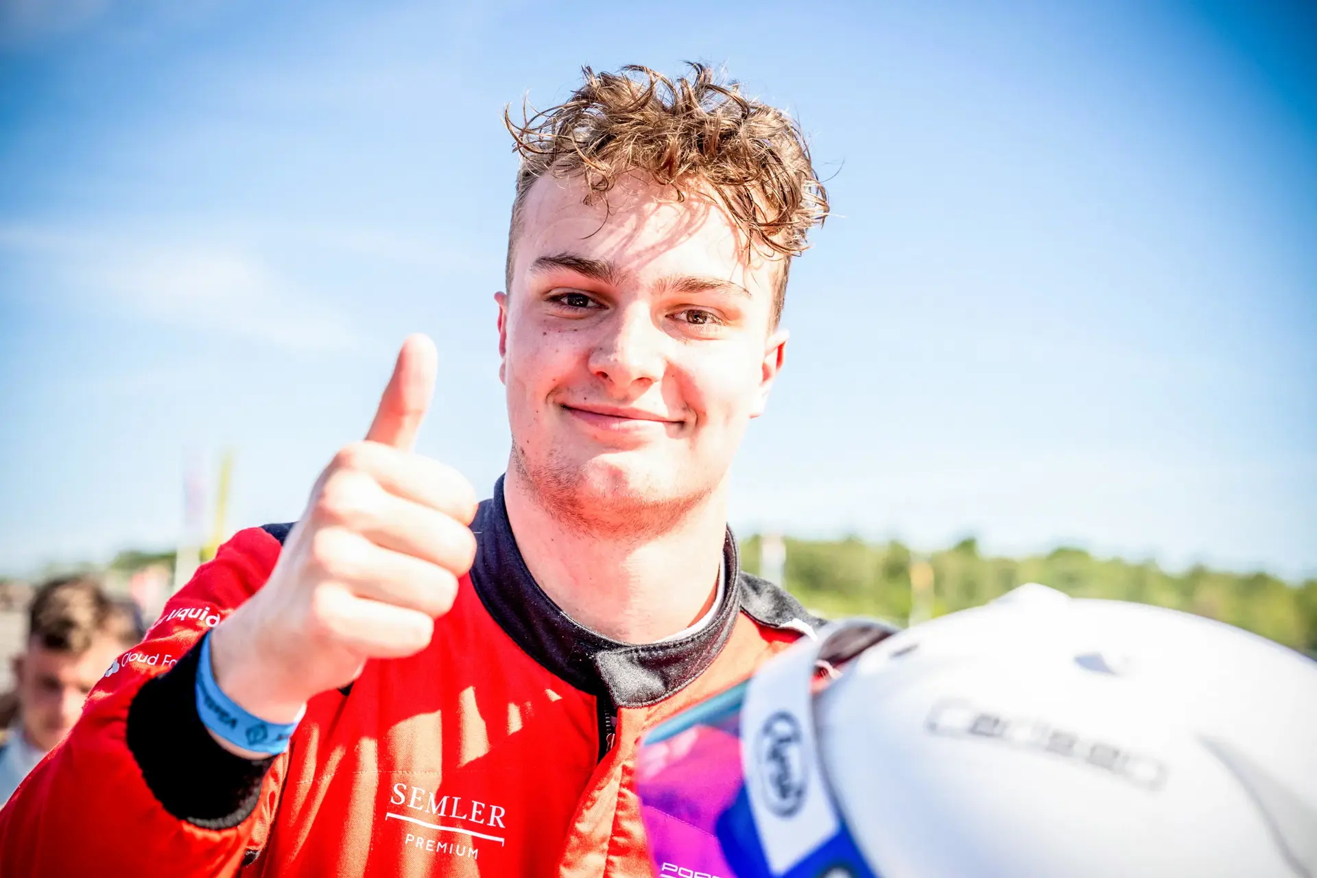 Racerkører i rød køredragt giver thumbs up ved banen på en solrig dag.