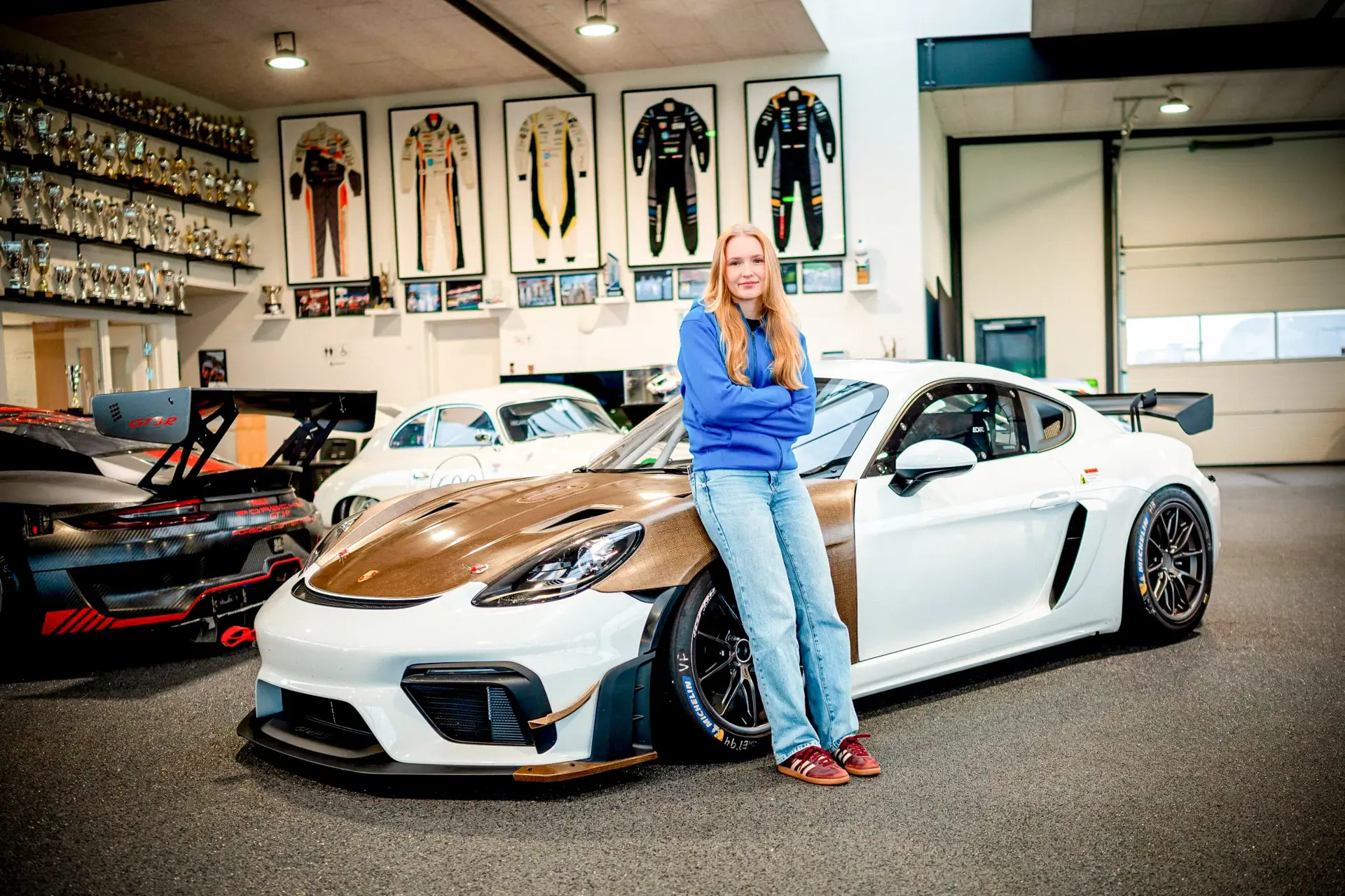 Ung kvinde står lænet op ad en hvid Porsche-racerbil i et værksted fyldt med pokaler og racerkombinationer på væggene.