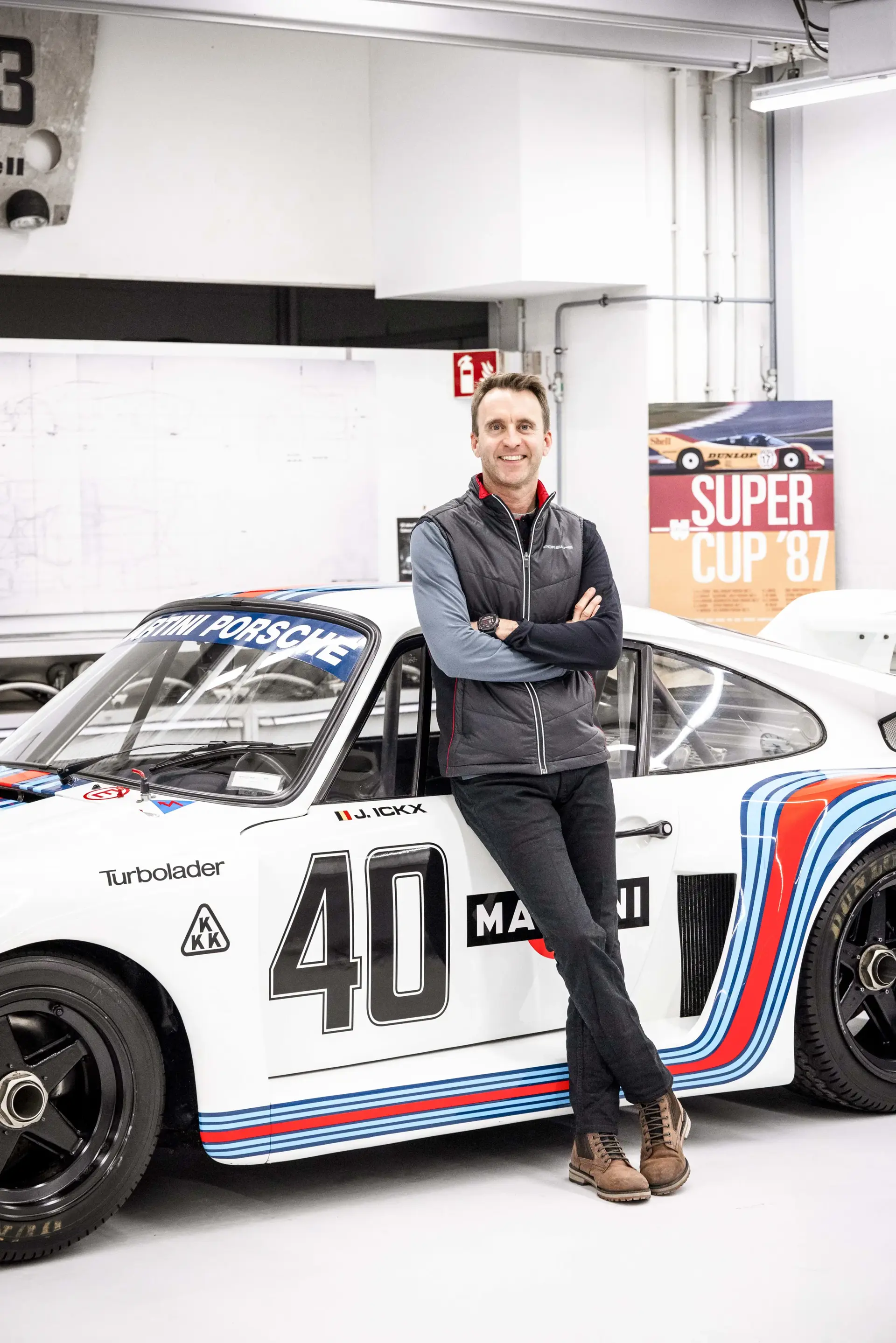 En person står lænet op ad en hvid klassisk Porsche-racerbil med Martini-bemaling i en lys garage.
